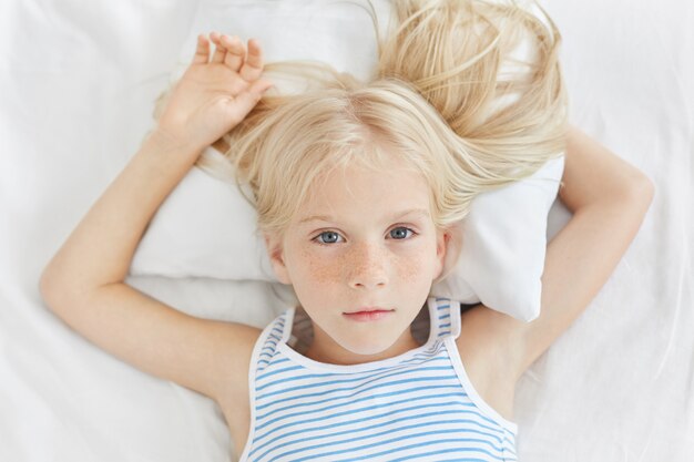 Entzückendes kleines Mädchen mit sommersprossiger Haut mit krankem Blick, während auf weißem Bett des Krankenhauses liegend, mit ihren blauen bezaubernden Augen schauend, Ruhe haben wollen.