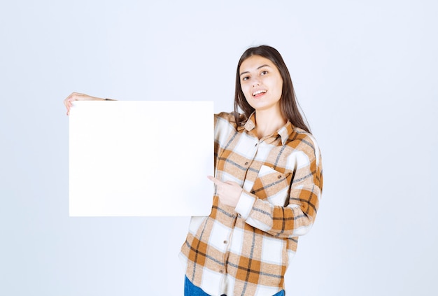 entzückendes Mädchen, das weiße große leere Karte auf weißer Wand zeigt.