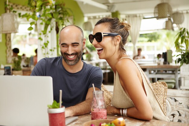 Entzückendes Paar, das laut lacht, während es mit seinem modernen Laptop im Straßencafé sitzt und Filme online mit kostenlosem Wi-Fi sieht.