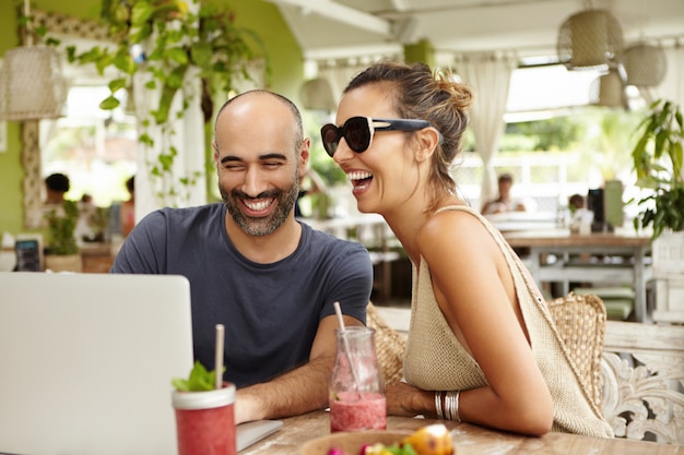 Entzückendes Paar, das laut lacht, während es mit seinem modernen Laptop im Straßencafé sitzt und Filme online mit kostenlosem Wi-Fi sieht.