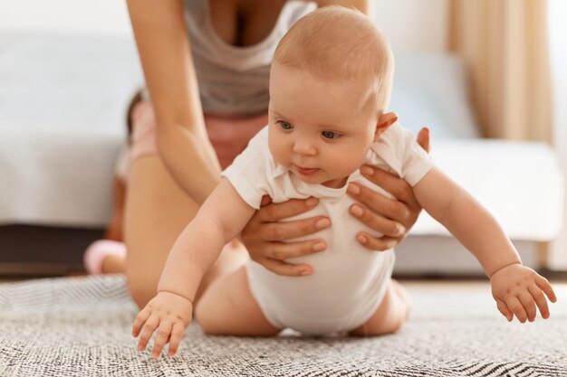 Entzückendes süßes Baby im weißen Body, das auf dem Teppich auf dem Boden kriecht, während die Mutter hilft und unterstützt, im hellen Raum zu Hause posiert, glückliche Kindheit.