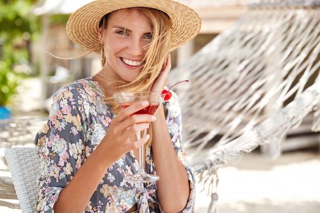 Entzückendes weibliches Modell mit attraktivem Blick trägt Strohhut und Hemd mit Blumendruck, trinkt erfrischenden Cocktail und ist mit gutem Resort zufrieden