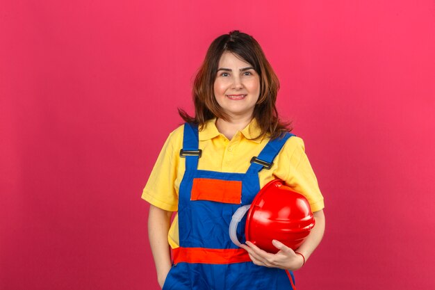 Erbauerfrau, die Bauuniform trägt, die Sicherheitshelm in der Hand mit Lächeln auf glücklichem Gesicht hält, das über isolierter rosa Wand steht