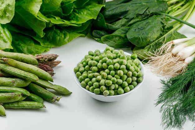 Erbsen in einer Schüssel mit grünen Schoten, Sauerampfer, Dill, Salat, Spargel, Frühlingszwiebeln hoher Winkelansicht auf einer weißen Wand