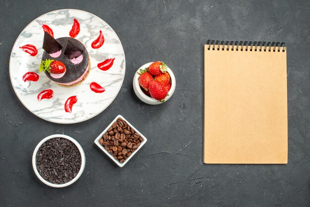 Erdbeer-Käsekuchen von oben auf weißen ovalen Tellerschalen mit Erdbeer-Schokoladen-Kaffeebohnen-Samen ein Notizbuch auf dunkler Oberfläche