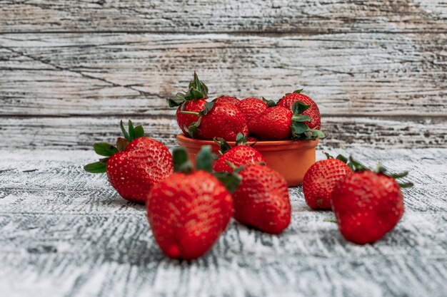 Erdbeeren der Seitenansicht in der braunen Schüssel mit mehreren Früchten um sie herum auf Stück Sack und grauem hölzernem Hintergrund. horizontaler Raum für Text