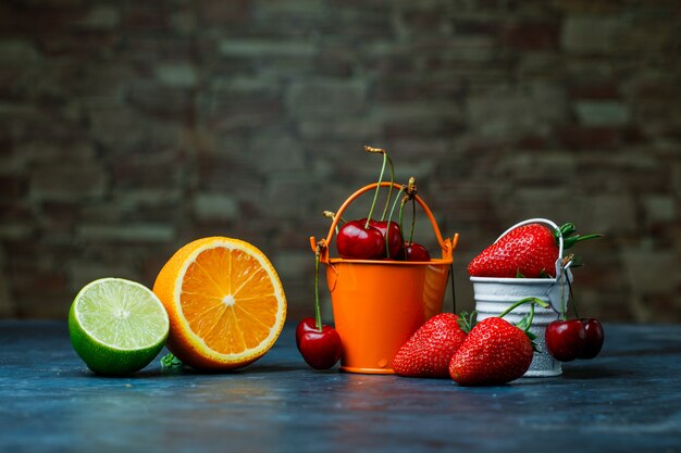 Erdbeeren und Kirschen in Mini-Eimern mit orange, Limetten-Seitenansicht auf Ziegelstein und blauem Hintergrund