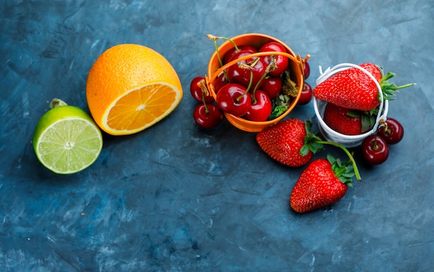 Kostenloses Foto erdbeeren und kirschen mit orange, limette in mini-eimern auf grungy blauem hintergrund, flach liegen.