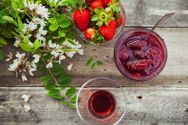 Erdbeermarmelade mit Löffel, ein Glas Tee, Erdbeeren, Pflanze in einem Teller auf Holztisch, Draufsicht.