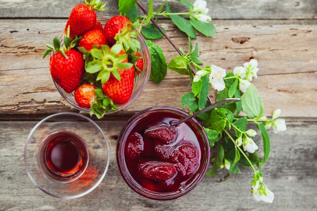 Erdbeermarmelade mit Löffel, Tee im Glas, Erdbeeren, Blumenzweig in einem Teller auf Holztisch, Draufsicht.