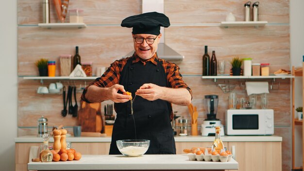 Erfahrener Bäcker, der Eier zum Backen mit Schürze knackt und Hobby genießt. Pensionierter älterer Koch mit Bonete, der von Hand mischt, in Glasschüssel Gebäckzutaten knetet und hausgemachten Kuchen zubereitet