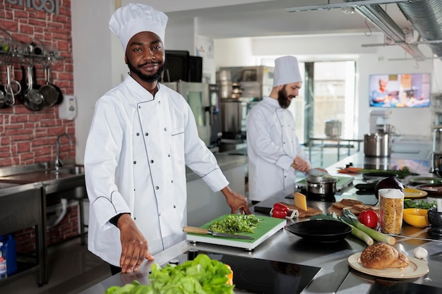 Erfahrener Küchenchef in Kochuniform bei der Zubereitung von Zutaten für das Gericht in der professionellen Küche des Restaurants. Afroamerikanischer Gastronomieexperte, der Vorbereitungsarbeiten für das Abendessen durchführt.