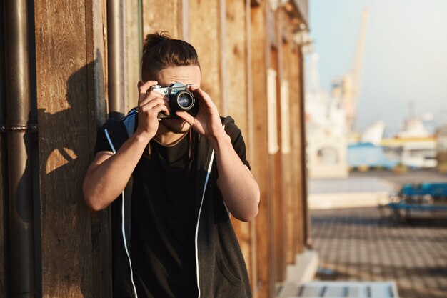 Erfassen Sie jeden Moment des Lebens. Außenaufnahme des jungen stilvollen Fotografen, der durch Weinlesekamera schaut, Aufnahmen des Hafens, der Yachten und der Küste macht und als freiberuflicher Kameramann arbeitet