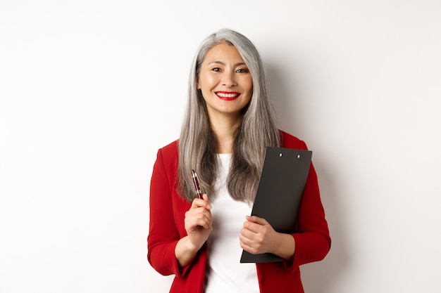 Erfolgreiche asiatische Chefin im roten Blazer, die Zwischenablage mit Dokumenten und Stift hält, arbeitet und glücklich aussieht, weißer Hintergrund.