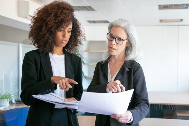 Erfolgreiche Büroarbeitgeber vergleichen Analysedaten miteinander. Konzentrierte selbstbewusste Managerinnen, die im Besprechungsraum auf Dokumente oder Berichte zeigen. Teamwork-, Geschäfts- und Managementkonzept