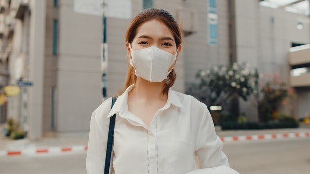 Erfolgreiche junge asiatische Geschäftsfrau in Modebürokleidung, die medizinische Gesichtsmaske trägt, die in der Straße lächelt