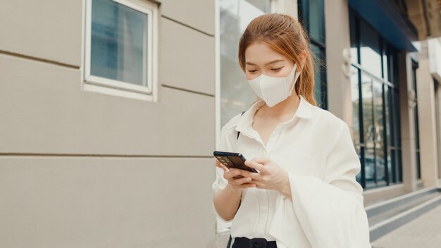 Erfolgreiche junge asiatische Geschäftsfrau in Modebürokleidung, die medizinische Gesichtsmaske unter Verwendung des Smartphones trägt