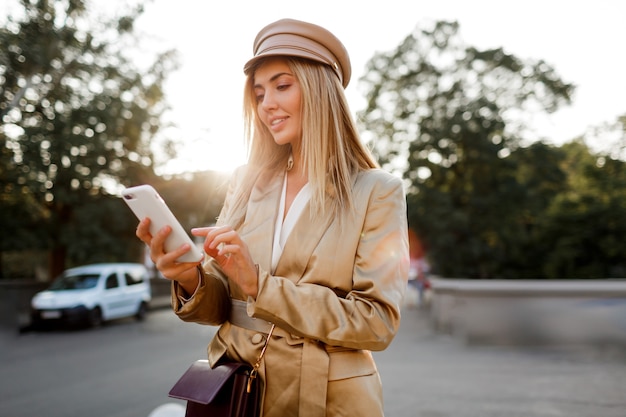 Erfolgreiche modische europäische Frau im eleganten lässigen Outfit, das mobyle Telefon im Freien aufwirft. Sonnenuntergangsfarben.