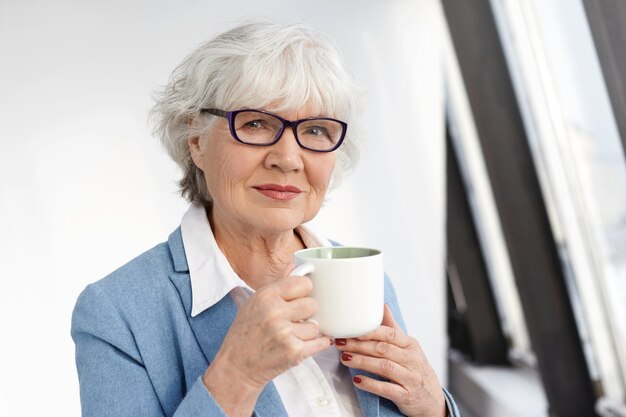 Erfolgreiche ordentliche kaukasische Geschäftsfrau mittleren Alters in formellen Kleidern und Brillen, die während der Kaffeepause Ruhe haben, Tasse halten und mit selbstbewusstem glücklichem Lächeln schauen. Menschen und Lebensstil