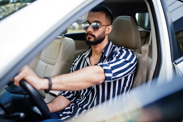 Erfolgreicher arabischer Mann in gestreiftem Hemd und Sonnenbrille posiert hinter dem Steuer seines weißen Geländewagens Stilvolle arabische Männer im Transport