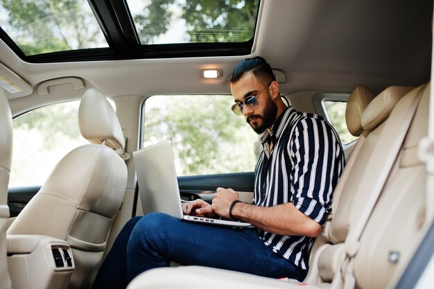 Erfolgreicher arabischer Mann in gestreiftem Hemd und Sonnenbrille posiert im weißen Geländewagen mit Laptop in den Händen