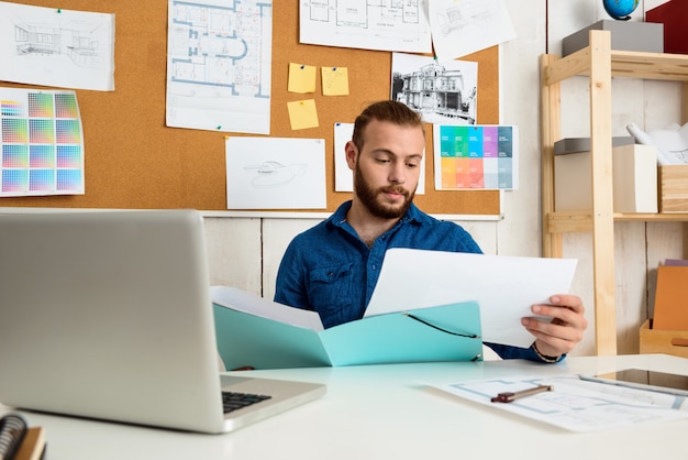 Erfolgreicher Geschäftsmann, der durch Papiere schaut und am Arbeitsplatz mit Laptop sitzt