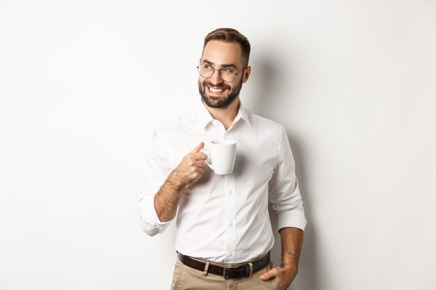 Erfolgreicher Geschäftsmann, der Kaffee trinkt, seitwärts mit zufriedenem Lächeln schaut und über weißem Hintergrund steht.