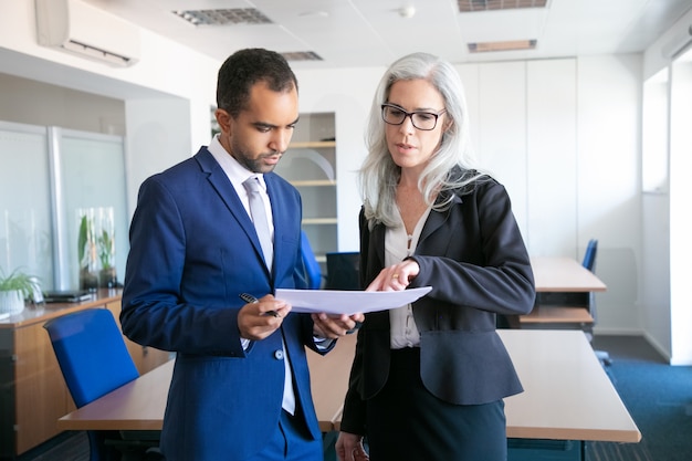 Erfolgreicher Geschäftsmann im Anzug, der Dokument für die Unterzeichnung liest, und weiblicher grauhaariger Manager in Brillen, die auf etwas im Bericht zeigen. Partner, die im Büro arbeiten. Geschäfts- und Managementkonzept