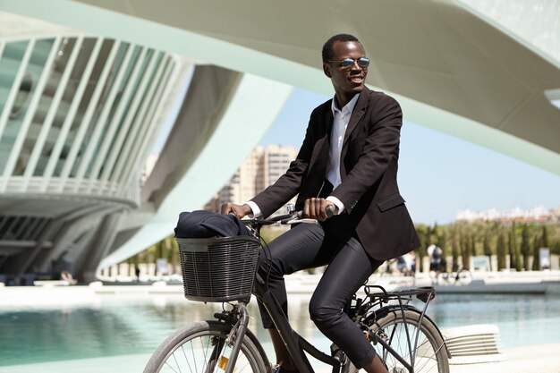 Erfolgreicher glücklicher afroamerikanischer Manager im schwarzen Anzug, der mit dem Fahrrad zum Büro pendelt. Dunkelhäutiger Angestellter, der sich beeilt, am Fahrrad zu arbeiten. Umweltfreundlicher Transport, urbaner Lebensstil und Transport