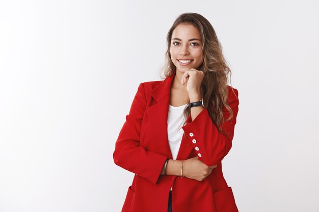 Erfolgreicher, gutaussehender Büromanager mit roter Jacke, der selbstsichere Pose mit Handkiefer hält, lächelt und sich sicher fühlt, ist ein Zeichen, steht selbstbewusst und arbeitet in einer gesunden Büroumgebung