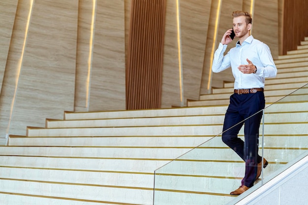Erfolgreicher, intelligenter kaukasischer Geschäftsmann, der auf der Treppe geht, lässt die Kommunikation mit dem Smartphone mit Glück und enthusiastischem, modernem Bürohintergrund kommunizieren