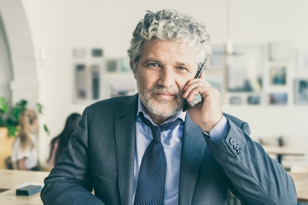 Kostenloses Foto erfolgreicher reifer geschäftsmann, der auf mobiltelefon spricht, bei der zusammenarbeit steht und sich auf schreibtisch schreibt