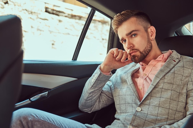 Erfolgreicher, stylischer junger Geschäftsmann in grauem Anzug und Ping-Shirt, der auf einem Rücksitz in einem Luxusauto fährt.