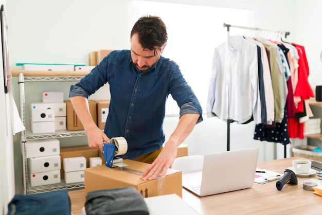 Erfolgreicher Unternehmer, der bereit ist, die Bestellungen der Kunden zu versenden. Geschäftsinhaber, der Klebeband auf ein großes Paket mit Bekleidungsprodukten legt