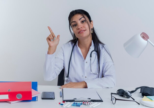 Erfreuliche junge ärztin, die medizinische robe und stethoskop trägt, die am schreibtisch mit medizinischen werkzeugen sitzen und hand auf schreibtisch setzen, die finger lokalisiert hebt