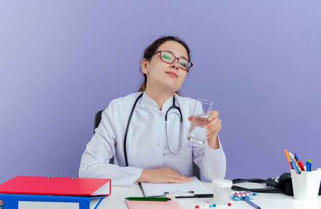 Erfreuliche junge Ärztin, die medizinische Robe und Stethoskop trägt, sitzt am Schreibtisch mit medizinischen Werkzeugen, die Glas des Wassers isoliert halten