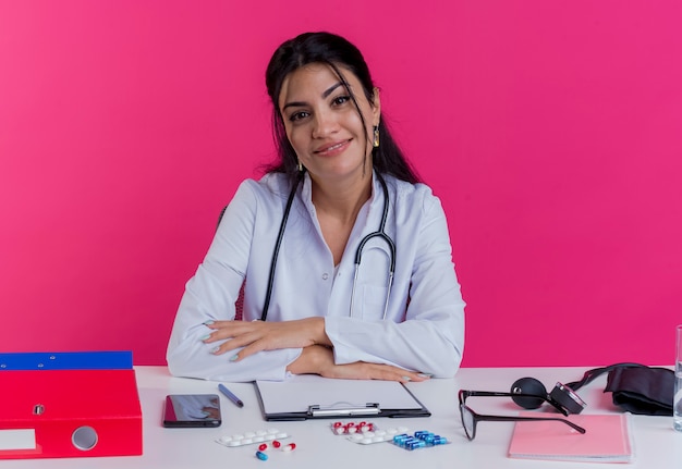 Erfreuliche junge Ärztin, die medizinische Robe und Stethoskop trägt, sitzt am Schreibtisch mit medizinischen Werkzeugen, die Hände auf Schreibtisch lokalisiert auf rosa Wand setzen