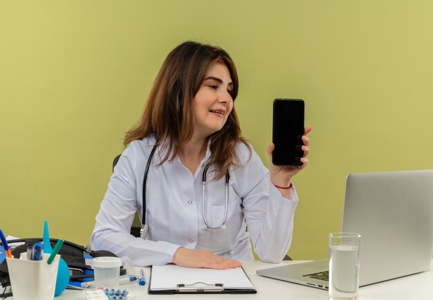 Erfreuliche Ärztin mittleren Alters, die medizinische Robe und Stethoskop trägt, die am Schreibtisch mit medizinischen Werkzeugen und Laptop sitzen und Handy lokalisiert betrachten