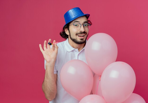 Erfreulicher gutaussehender Mann, der Brille und blauen Hut hält, der Ballone und Pfeife lokalisiert auf rosa Hintergrund hält