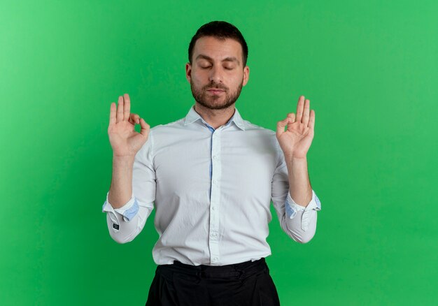Erfreulicher gutaussehender Mann gibt vor, mit geschlossenen Augen zu meditieren, die auf grüner Wand isoliert werden