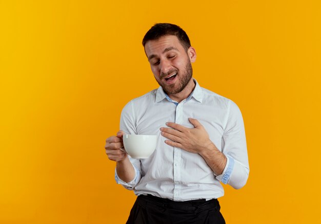 Erfreulicher gutaussehender Mann hält Tasse legt Hand auf Brust isoliert auf orange Wand