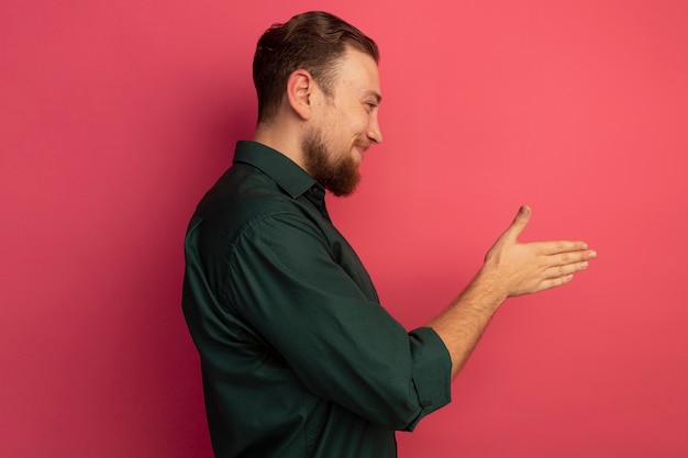 Erfreulicher hübscher blonder Mann steht seitlich und hält Hand lokalisiert auf rosa Wand heraus