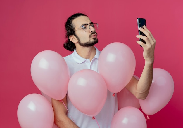 Erfreulicher hübscher Mann, der Brillen hält, die Luftballons halten und ein Selfie lokalisiert auf rosa Hintergrund nehmen