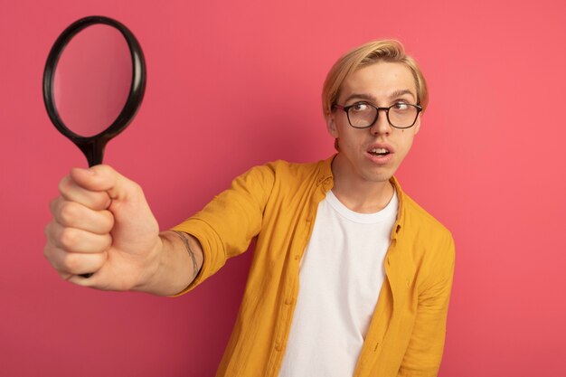 Erfreulicher junger blonder Kerl, der gelbes T-Shirt und Brille hält, die Lupe hält, die auf Rosa lokalisiert wird