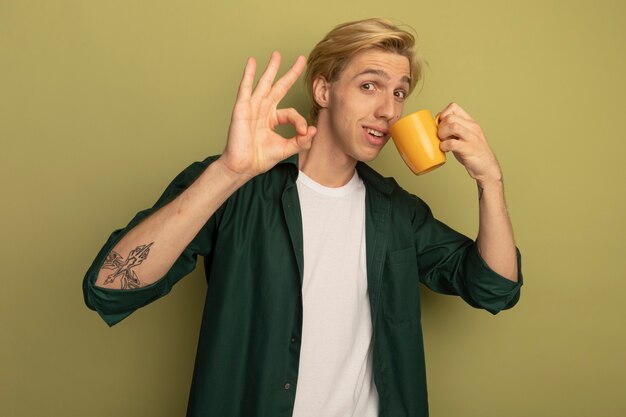 Erfreulicher junger blonder Kerl, der grünes T-Shirt trägt, trinkt Tee und zeigt okay Geste