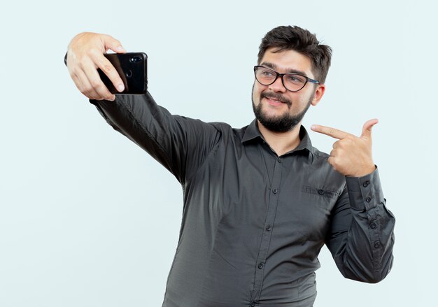 Erfreulicher junger Geschäftsmann, der Brille trägt, macht ein Selfie und zeigt isoliert auf weißem Hintergrund auf sich