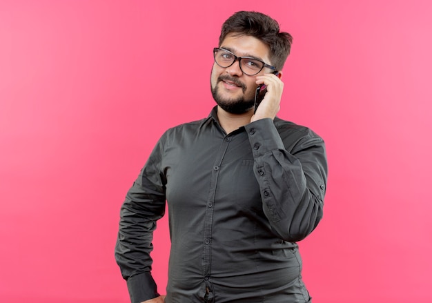 Erfreulicher junger geschäftsmann, der brille trägt, spricht am telefon und legt hand auf hüfte lokalisiert auf rosa hintergrund