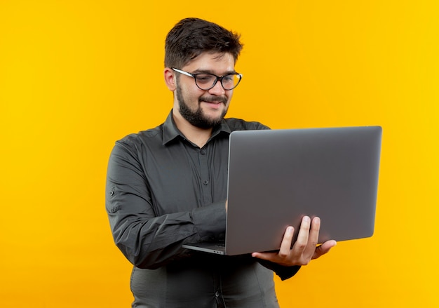 Erfreulicher junger Geschäftsmann, der Brillen hält und gebrauchten Laptop lokalisiert auf gelbem Hintergrund trägt