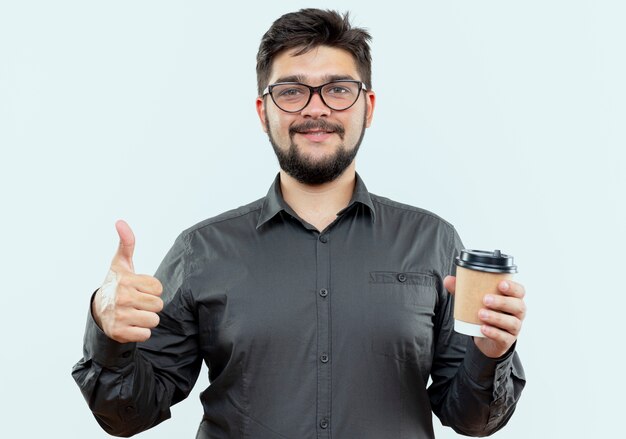 Erfreulicher junger Geschäftsmann, der Brillen trägt, die Tasse Kaffee seinen Daumen oben auf weißem Hintergrund halten