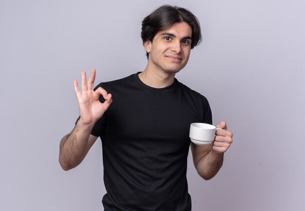 Erfreulicher junger gutaussehender Mann, der schwarzes T-Shirt hält, das Tasse Kaffee hält und okay Geste auf weißer Wand isoliert zeigt